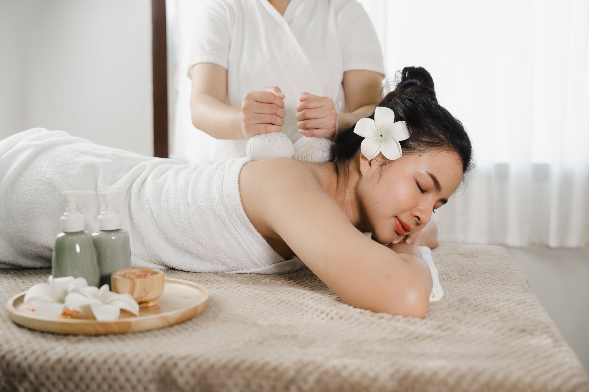 Relaxed pretty asian young woman enjoying remedial body massage done by professional masseur in spa room, with herbal compress balls in therapy spa salon.