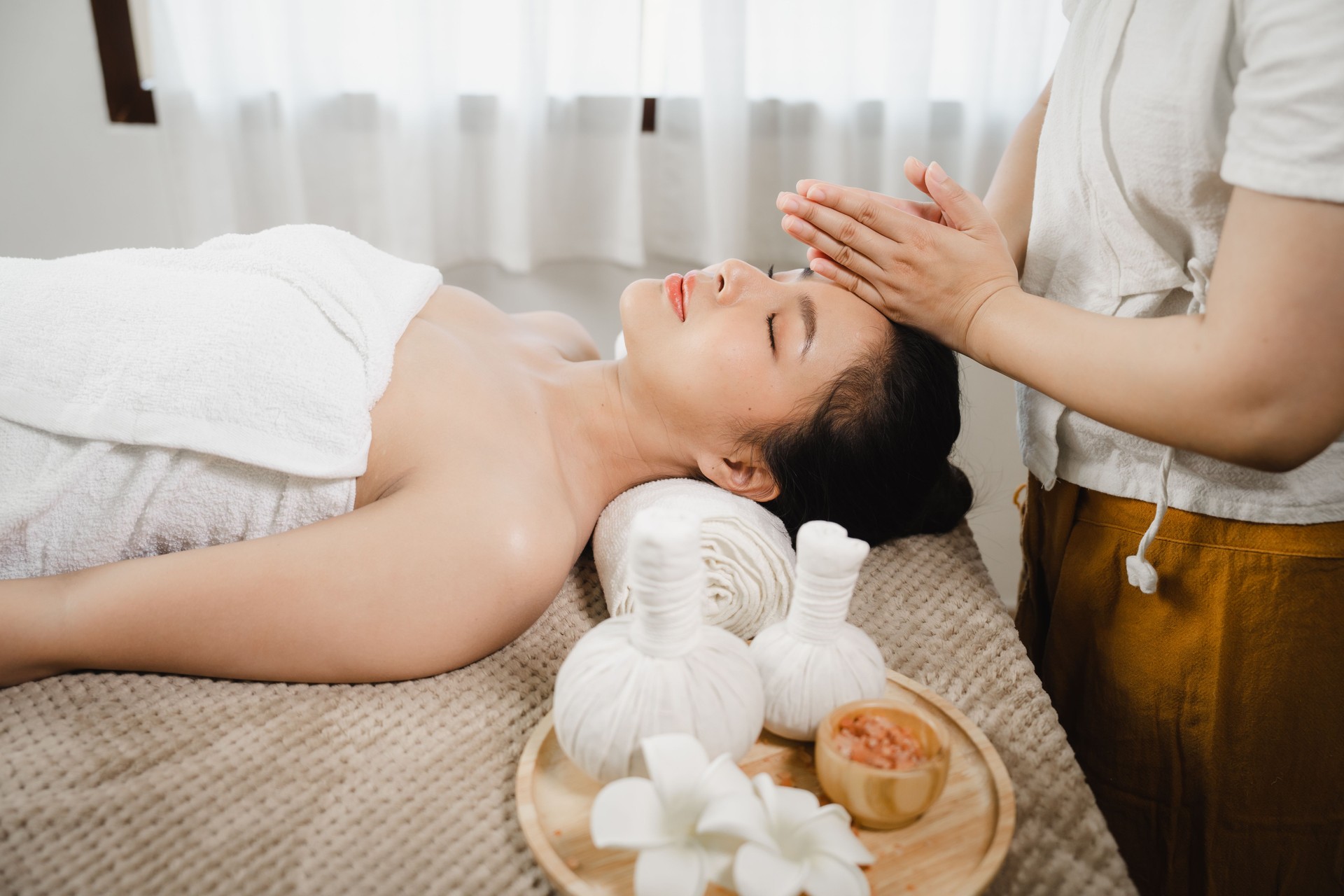 Pretty asian woman is given rejuvenating facial massage in an aroma room. Female therapist massaging the face and forehead of a relaxed client to a young woman. Alternative medicine.